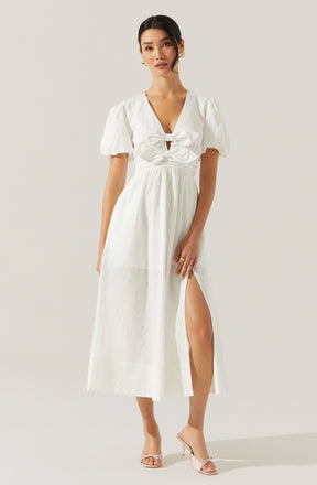 A woman is wearing a white maxi dress with short, puffed sleeves and a deep V neckline. The dress features a ruched bodice with a large bow at the front and a high slit on one side, revealing her leg. She is also wearing clear, open-toe high heels.