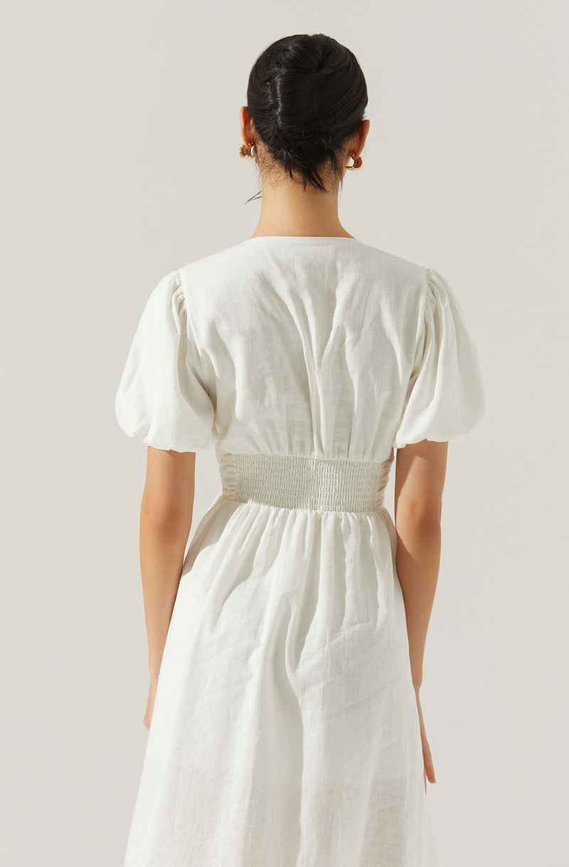 A woman is wearing a white dress with puffed short sleeves, a fitted waistband, and a gathered skirt. The dress appears to be made of a light, airy fabric, and the back features a central seam running vertically. The waistband is wide and elastic, creating a cinched waist effect. The woman has dark hair styled in an elegant updo and is accessorized with earrings.