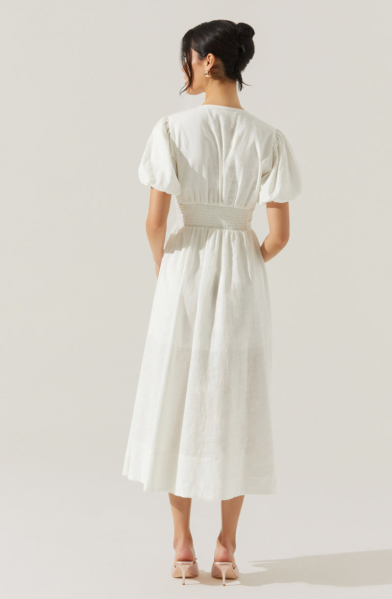 A woman is wearing a long, white dress with puffed short sleeves, a fitted waist with gathered detailing, and a full-length skirt that reaches mid-calf. She is standing with her back to the camera, and her hair is styled in an elegant updo. She is also wearing light-colored, open-toe high heels.