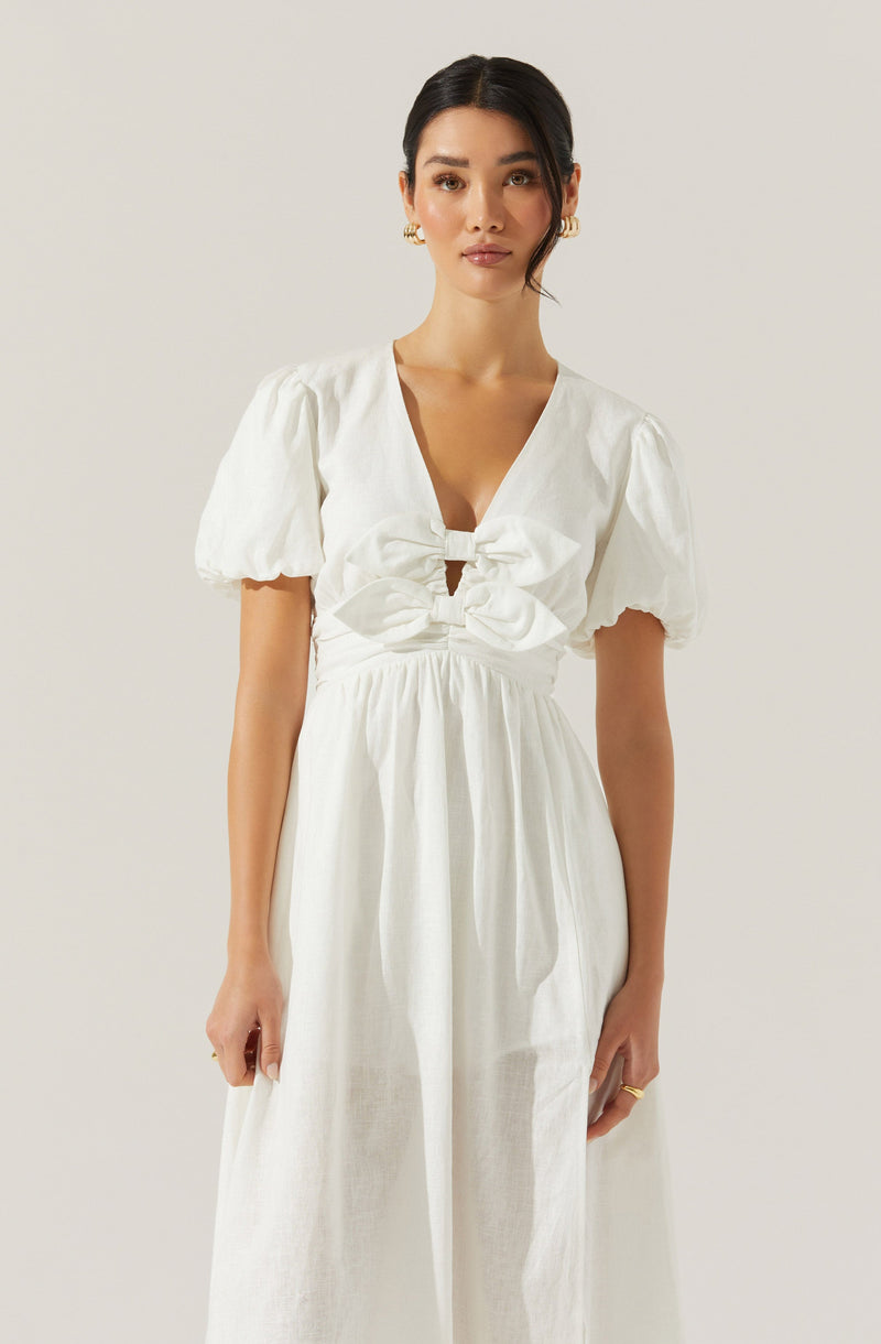A woman is wearing a white dress with short, puffed sleeves and a deep V neckline. The dress features a large bow detail at the bust, gathered fabric at the waist, and a flowing skirt. She has dark hair styled in a low bun and is accessorized with gold hoop earrings and a ring.