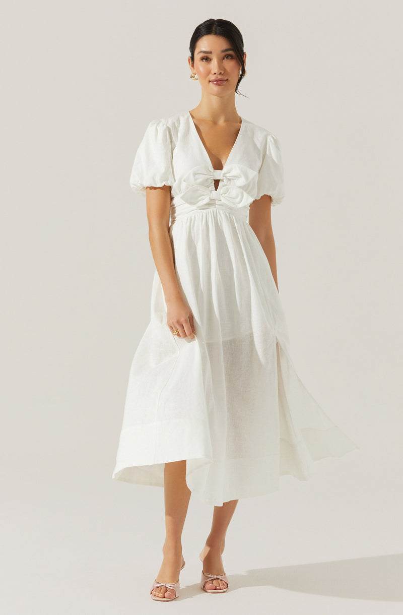 A woman is wearing a white dress with puffed short sleeves, a V-neckline, and a large decorative bow at the front of the waist. The skirt is flowy and midi-length, made of a lightweight, slightly textured fabric. She is also wearing light-colored, open-toe heels with a bow detail at the front.