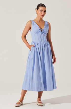 A woman is wearing a light blue sleeveless dress with a fitted bodice featuring a lace-up detail at the front. The dress has a full, gathered skirt that reaches mid-calf length. She completes the look with white, closed-toe shoes that have a small heel.