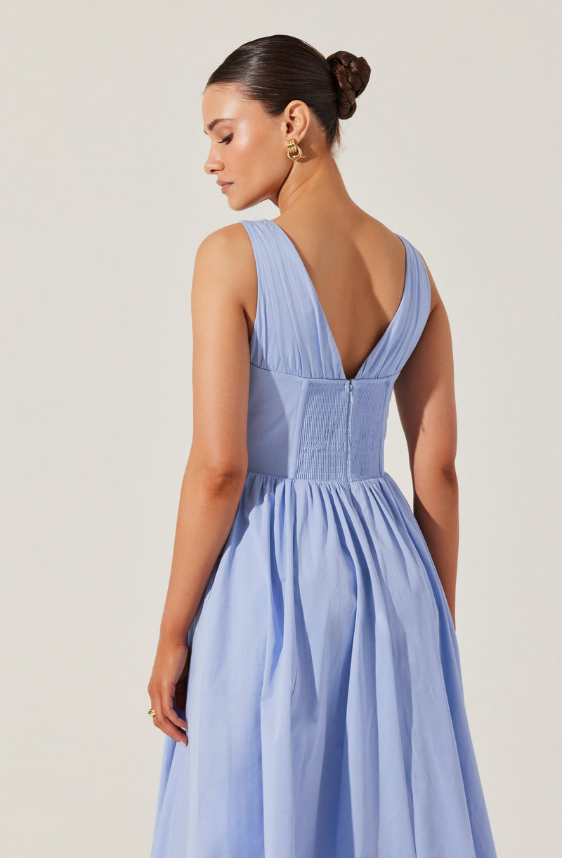 A woman is wearing a sleeveless, light blue dress with a deep V back. The dress features gathered fabric on the shoulders, a fitted bodice with pleats and smocking detail at the waist, and a full, flowing skirt with pleats. She has dark hair styled in a neat bun and is accessorized with gold hoop earrings.