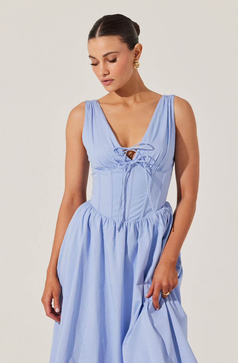 A woman is wearing a sleeveless, light blue dress with a deep v-neckline and gathered fabric at the shoulders. The bodice features a lace-up detail at the front with thin ties, and the waist is fitted, flowing into a full, gathered skirt. The fabric appears lightweight and has a slight sheen. She has dark hair styled in a sleek updo, accessorized with gold, hoop earrings.