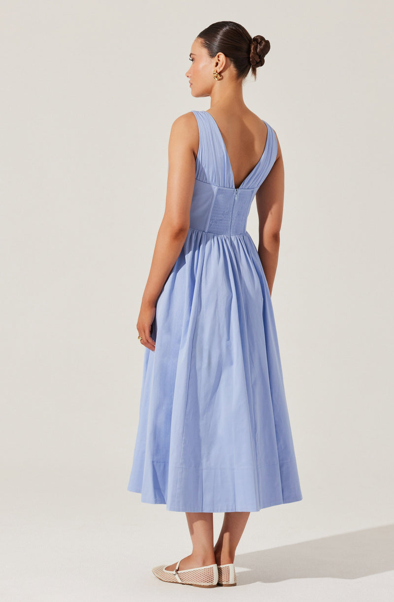 A woman is wearing a sleeveless, light blue, midi-length dress with a deep V neckline in the back. The dress features a fitted bodice that zips up the back and a gathered, flowing skirt. She is also wearing open-toe, beige shoes with a low heel.