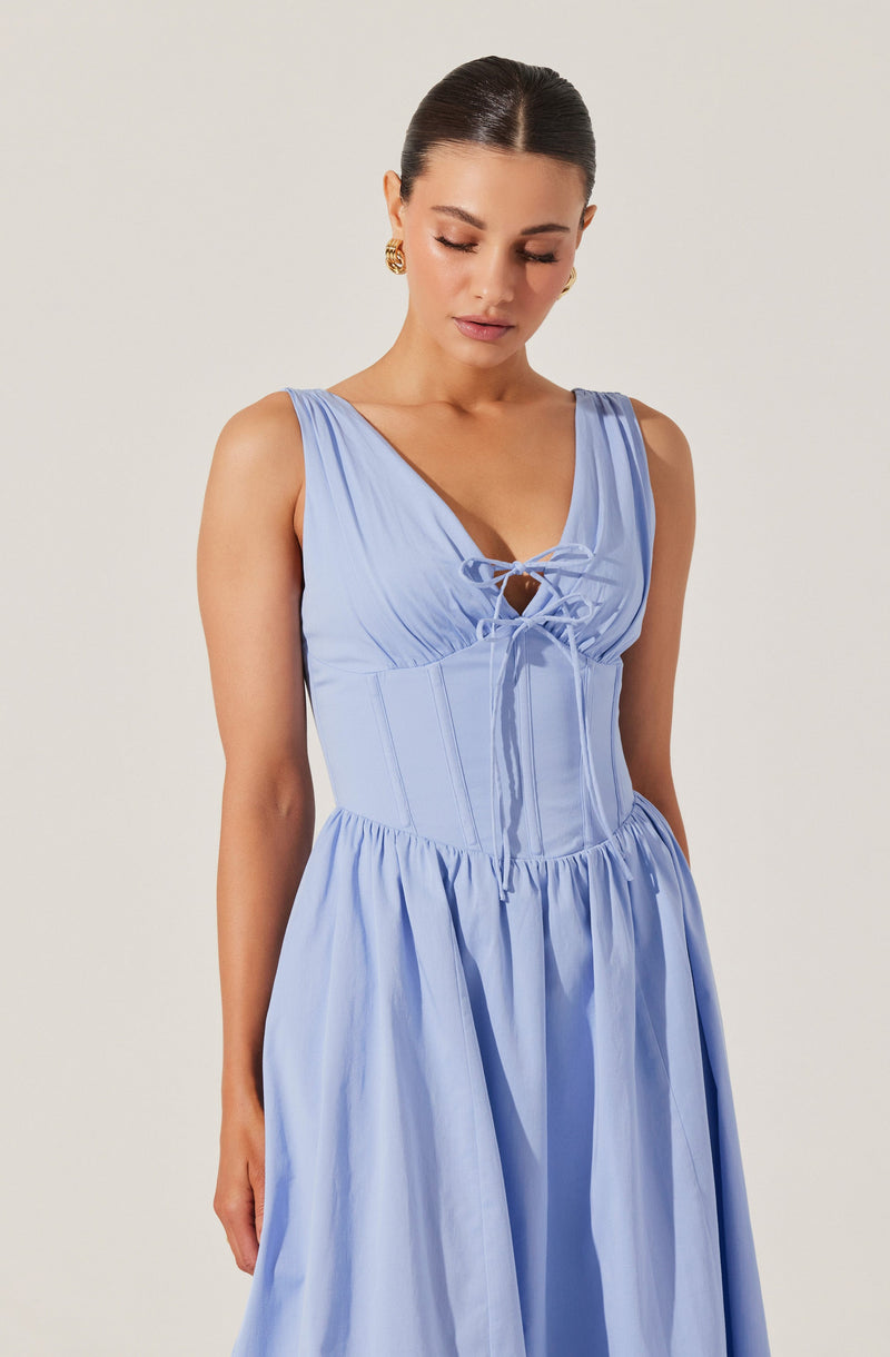 A woman is wearing a sleeveless, light blue dress with a fitted bodice featuring vertical seam details and a lace-up neckline with a small keyhole opening. The dress has a full, gathered skirt that falls to a midi or ankle length. She is accessorized with gold earrings and has her dark hair styled in a sleek, low bun.