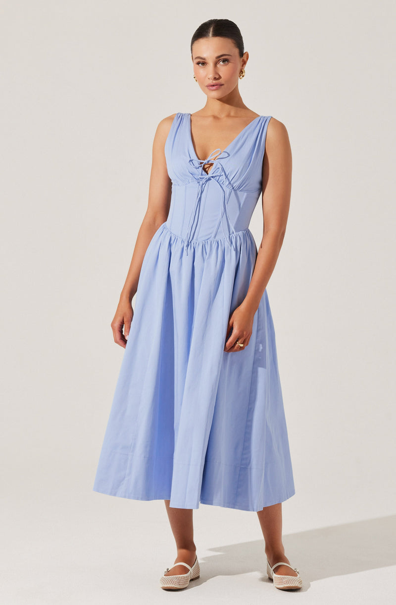 A woman is wearing a sleeveless, light blue dress with a lace-up detail at the front neckline. The dress features a fitted bodice with vertical seam accents and a gathered, voluminous skirt that falls to mid-calf length. She is also wearing beige, woven, closed-toe shoes and small, gold earrings.