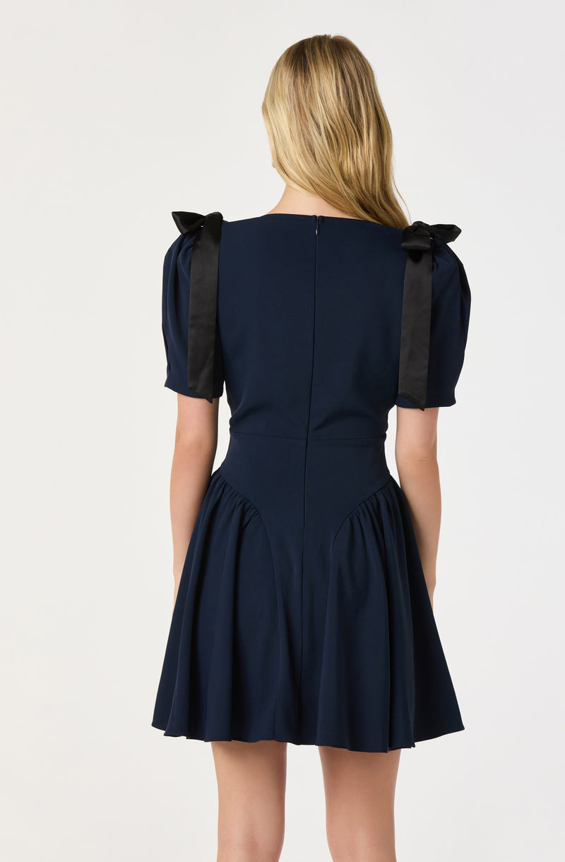 A woman is wearing a navy blue dress with short puffed sleeves that have black ribbon bows tied at the shoulders. The dress has a fitted bodice and a gathered A-line skirt, with a visible zipper running down the back. The fabric appears to be smooth and the dress has a classic, elegant style.