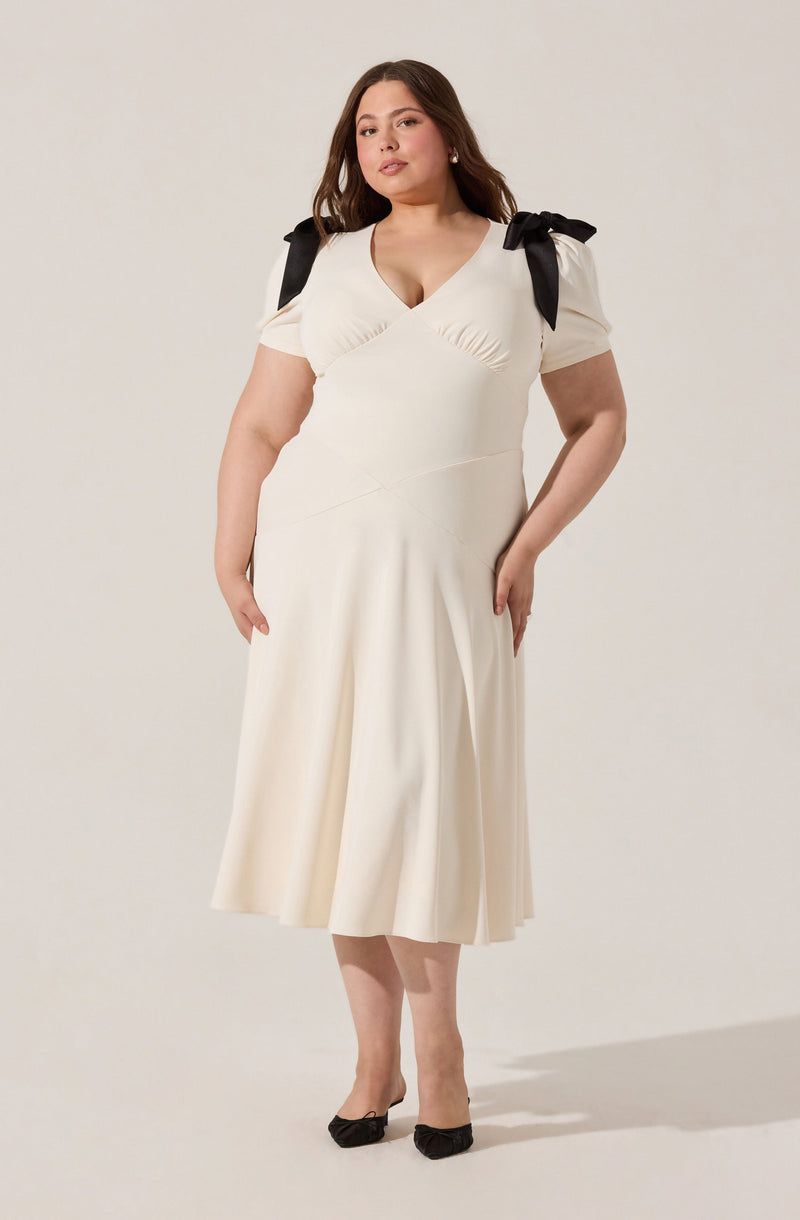 A woman is wearing a cream-colored, midi-length dress with a fitted bodice and a flared skirt. The dress features short puff sleeves with black ribbon bows on each shoulder. The neckline is a V-shape with subtle ruching details.