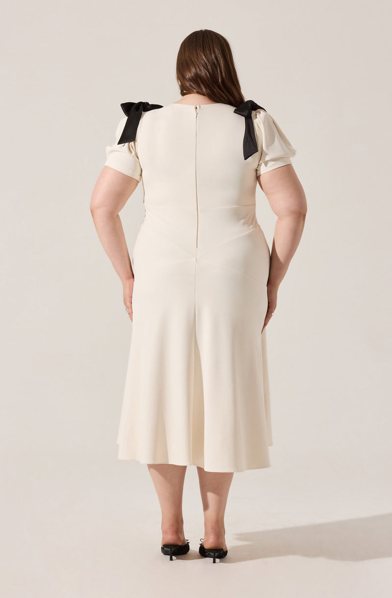 A woman is wearing a cream-colored dress with short puffed sleeves that have black bows on the shoulders. The dress has a fitted waist and a back zipper, with pleats in the skirt that fall to mid-calf length. She is wearing black open-toe heels.