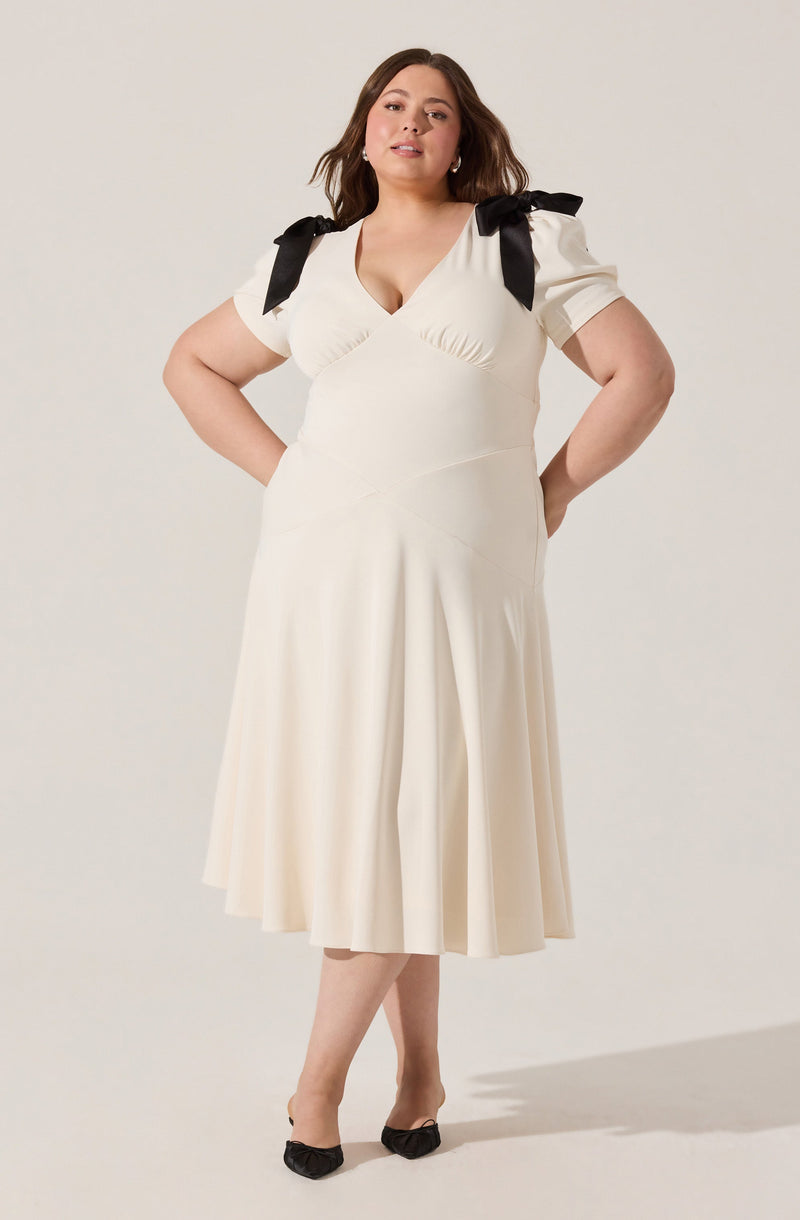 A woman is wearing a cream-colored dress with a V-neckline and short puff sleeves. The dress features black ribbon bows on each shoulder, tied into large bows, adding a decorative touch. She is also wearing black shoes with a bow detail on the front.