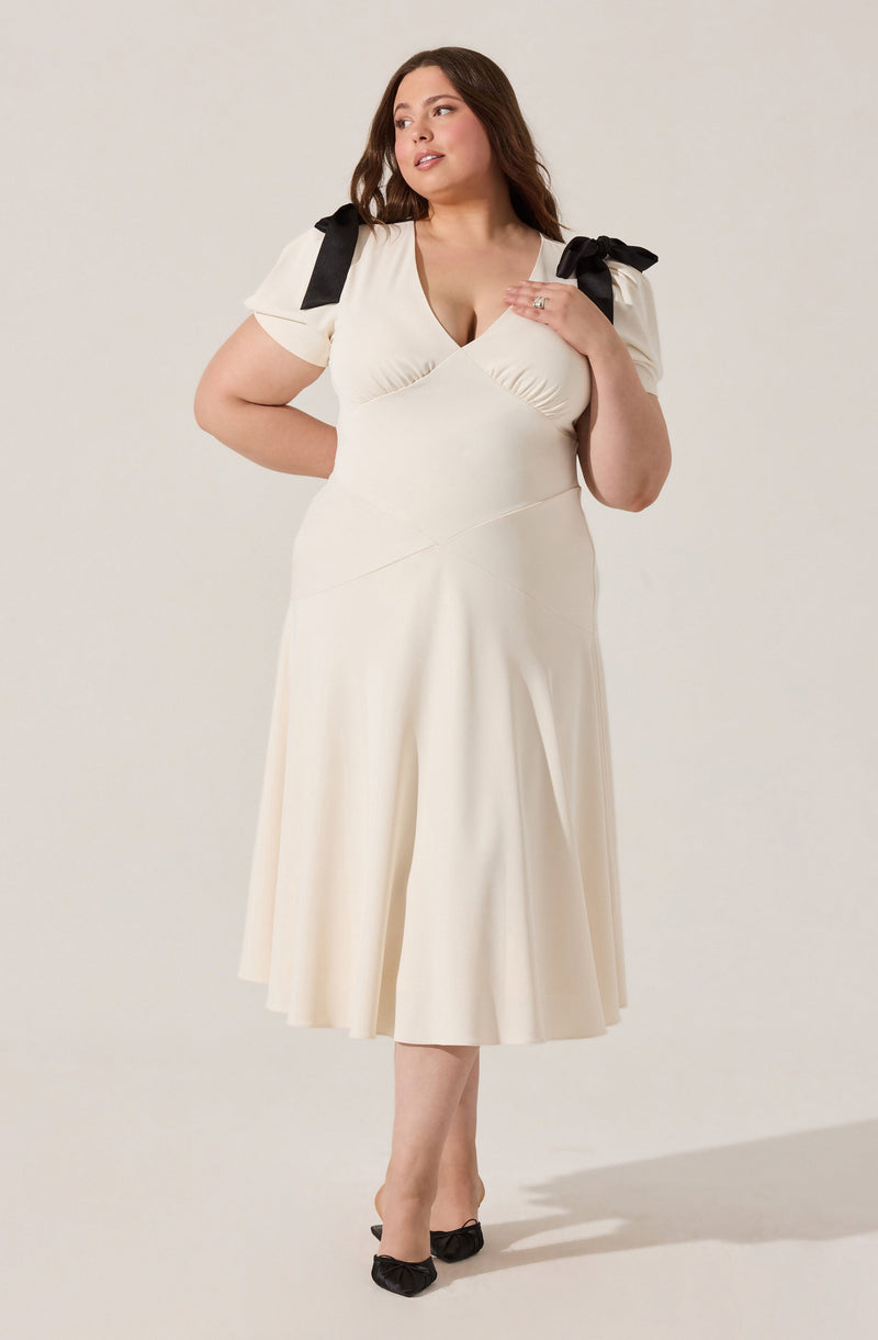 A woman is wearing a cream-colored dress with short sleeves and a V-neckline. The dress features black bows on the shoulders and has a fitted bodice with gathered detailing at the bust. The skirt of the dress is A-line, midi-length, and flows slightly away from the waist. She is also wearing black shoes with bows at the toes.