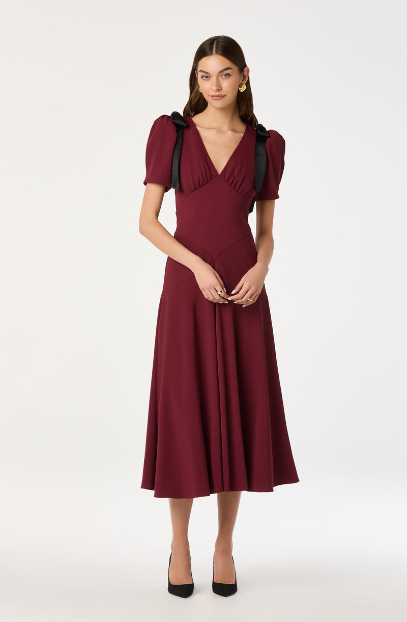 A woman is wearing a burgundy midi dress with a fitted bodice and a full, pleated skirt that falls below her knees. The dress features short puffed sleeves with black bows on each shoulder and a deep V-neckline. She is also wearing black high heels and large gold earrings.