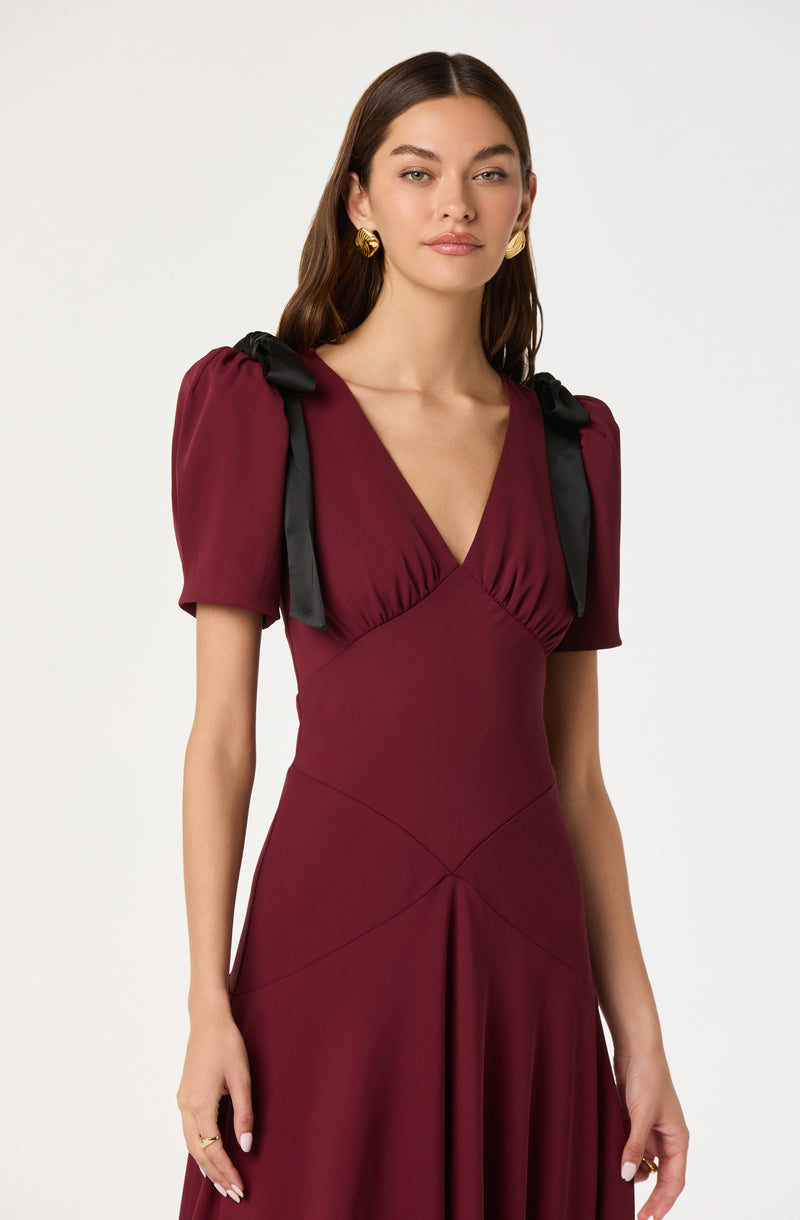 A woman is wearing a deep burgundy dress with a fitted bodice and a V-neckline. The dress features short puffed sleeves that are gathered at the shoulders and tied with black satin ribbons. The skirt has a flowing silhouette with seam details that create a structured look. She accessorizes with gold earrings and a simple ring.