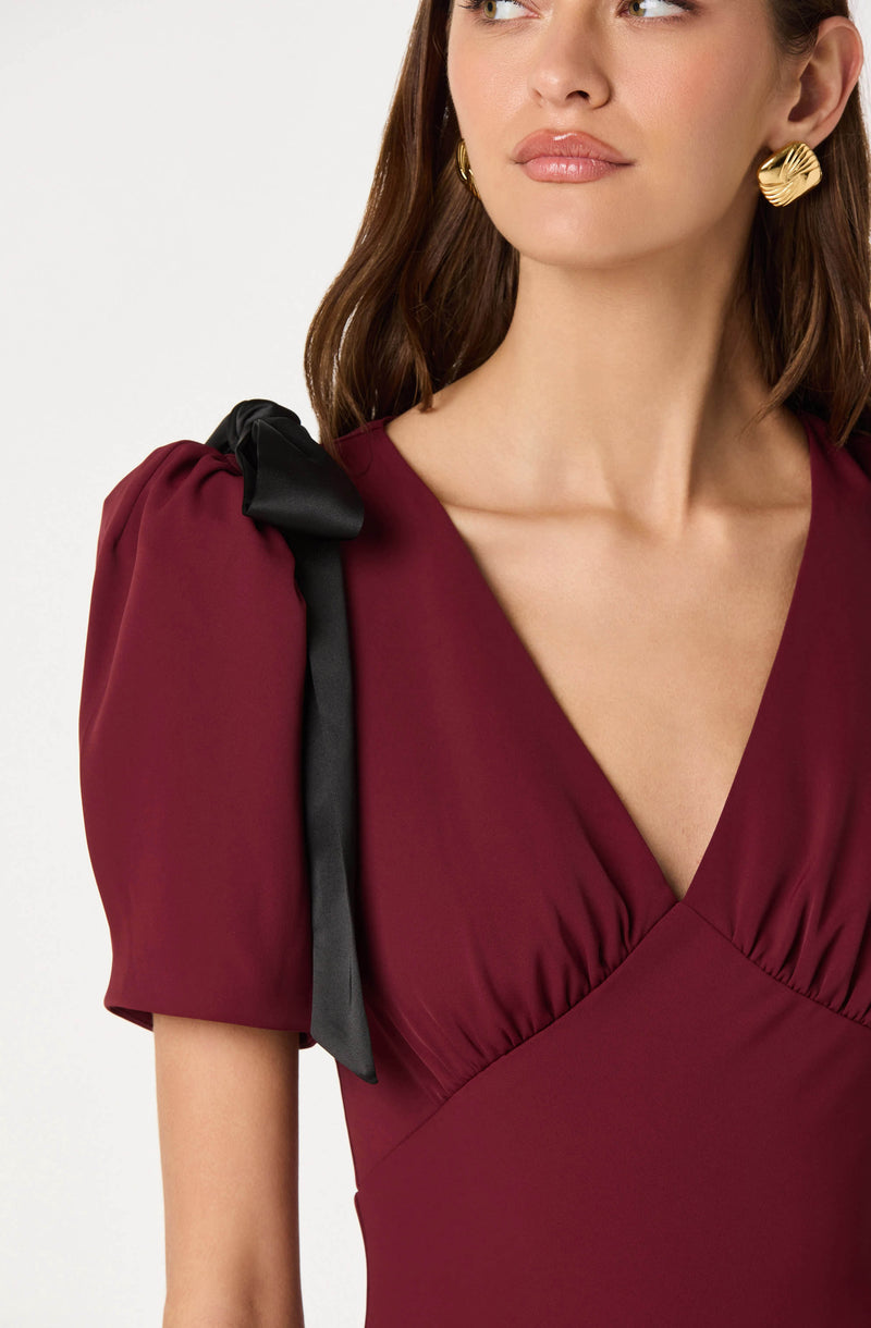 A woman is wearing a deep burgundy dress with puffed short sleeves. The shoulder of the dress has a black ribbon tied into a bow. She is accessorized with large, gold, textured hoop earrings.