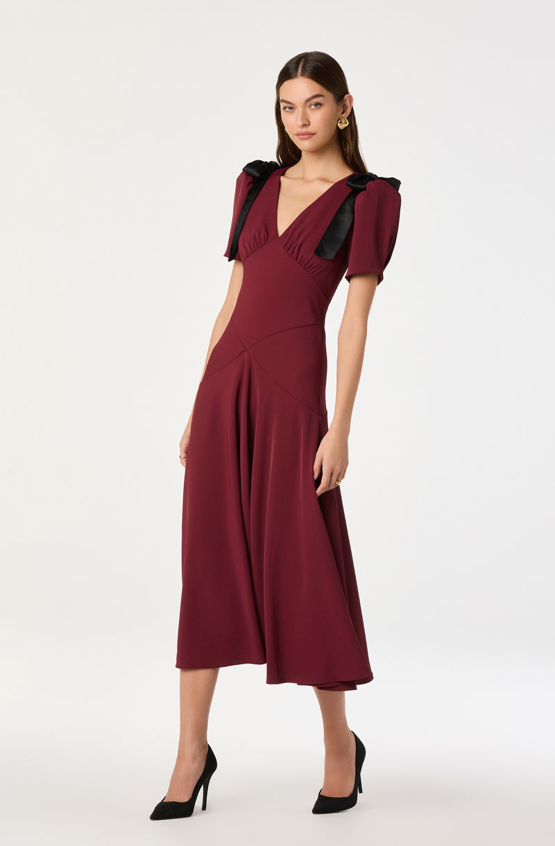 A woman is wearing a deep red, fitted midi dress with short puff sleeves and a V-neckline. The dress has a gathered detail at the front and a slightly flared hem. She is accessorizing with gold earrings and black high-heeled shoes. She also has black ribbons tied over her shoulders.