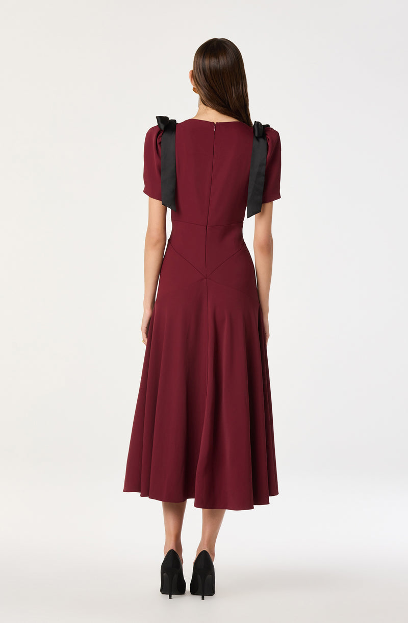 A woman is wearing a long, burgundy dress with a fitted bodice and puffed short sleeves. The dress has a flared, pleated skirt that reaches mid-calf length. She has black ribbon bows on her shoulders, and she is wearing black high-heeled shoes.