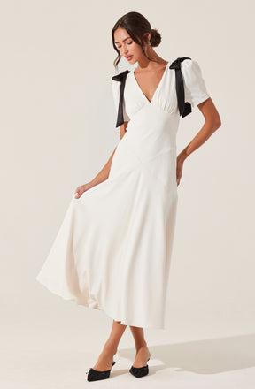 A woman is wearing a white, ankle-length dress with a V-neckline. The dress has puffed short sleeves with black bows on the shoulders. She is also wearing black pointed-toe shoes with small bows on the front.