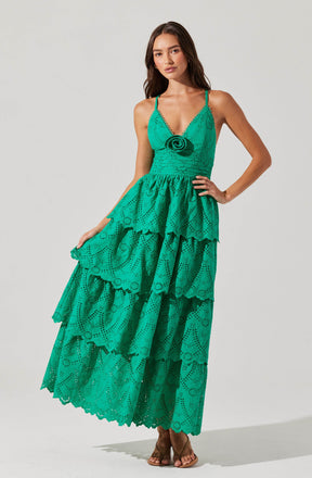 A woman is wearing a vibrant, turquoise, eyelet maxi dress with tiered ruffles. The dress features a fitted bodice with thin spaghetti straps, and a decorative rosette detail at the front. The skirt has multiple layers with scalloped edges, creating a flowing, tiered silhouette.