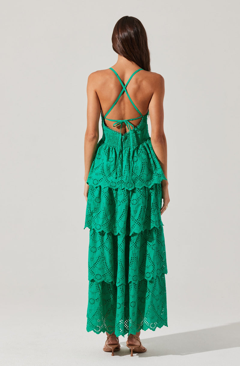 A woman is wearing a long, flowing, green dress with multiple tiers. The dress features eyelet embroidery and ruffled layers throughout the length. The back of the dress has thin, crisscrossing spaghetti straps that tie at the waist, revealing her back.