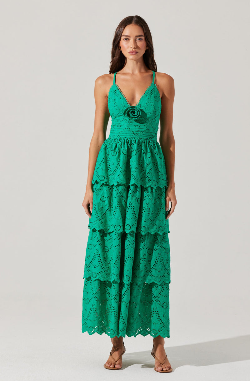 A woman is wearing a long, bright turquoise dress made of eyelet fabric with intricate cut-out patterns. The dress features a fitted bodice with adjustable spaghetti straps, a decorative rosette detail at the center of the bust, and a tiered, flowing skirt with multiple ruffled layers that extend to the floor.