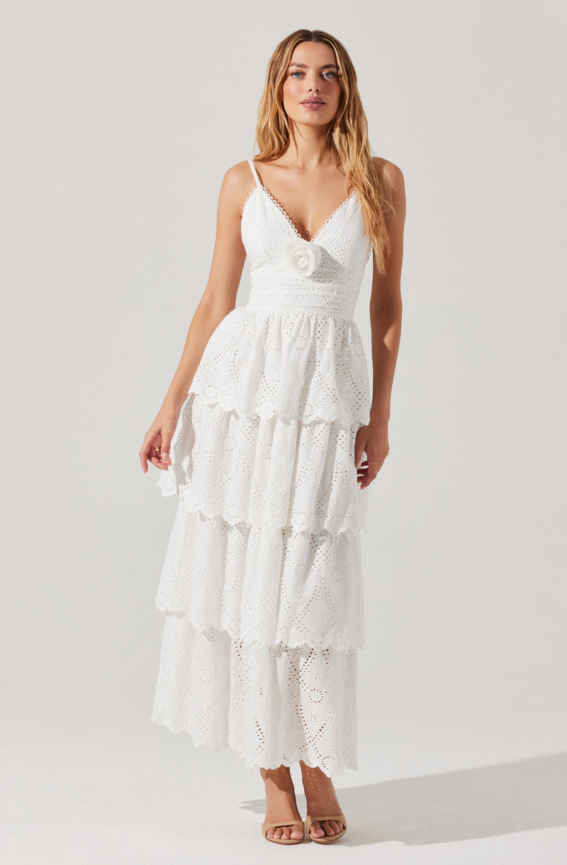 The woman is wearing a white, tiered, eyelet maxi dress with a fitted bodice and thin spaghetti straps. The dress features a decorative flower detail at the center of the bust and has a ruffled, layered skirt. She is also wearing open-toed, nude high-heeled sandals.