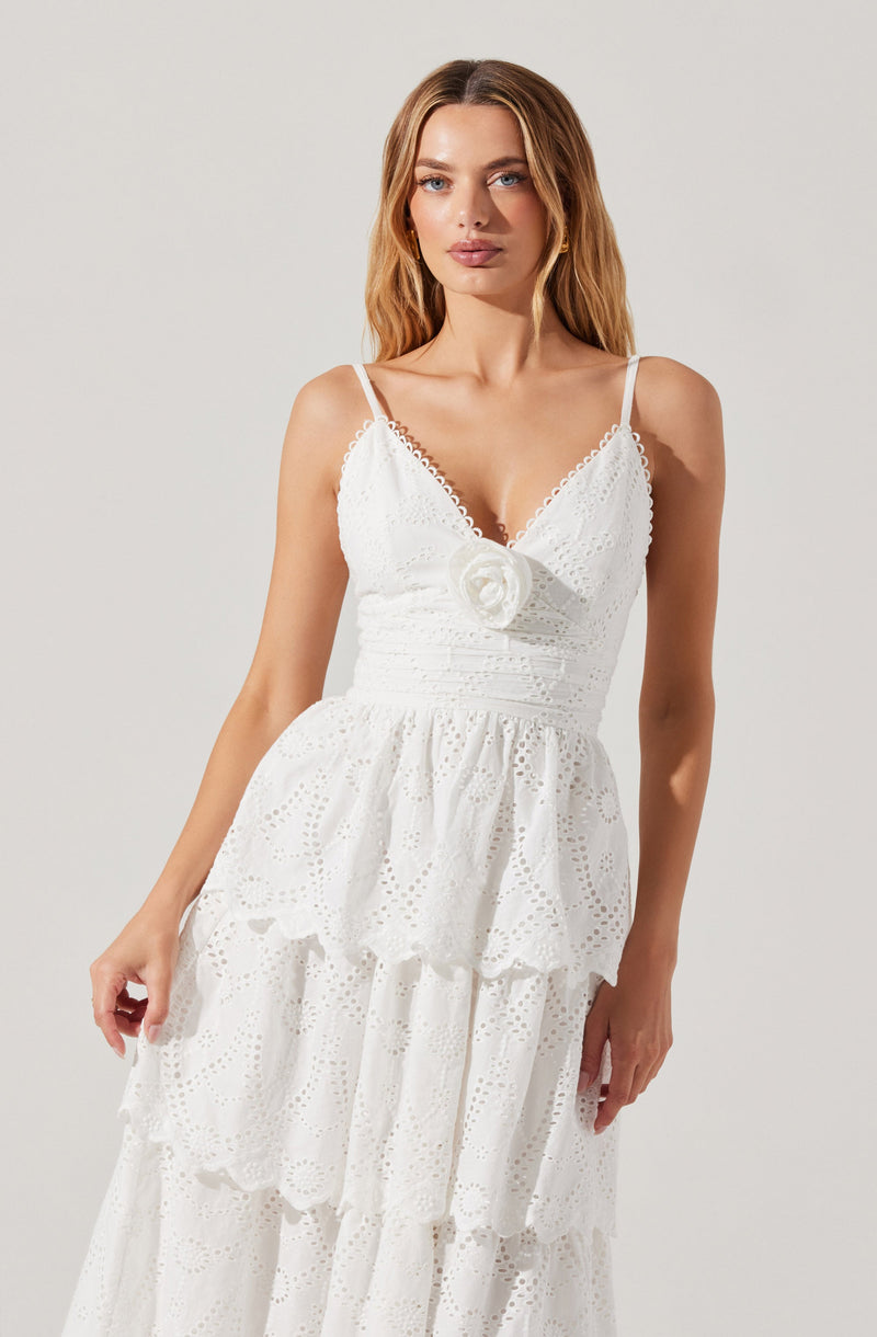 A woman is wearing a white dress with thin spaghetti straps. The dress features intricate eyelet embroidery and tiered ruffles on the skirt. There is a decorative fabric flower at the center of the bust.