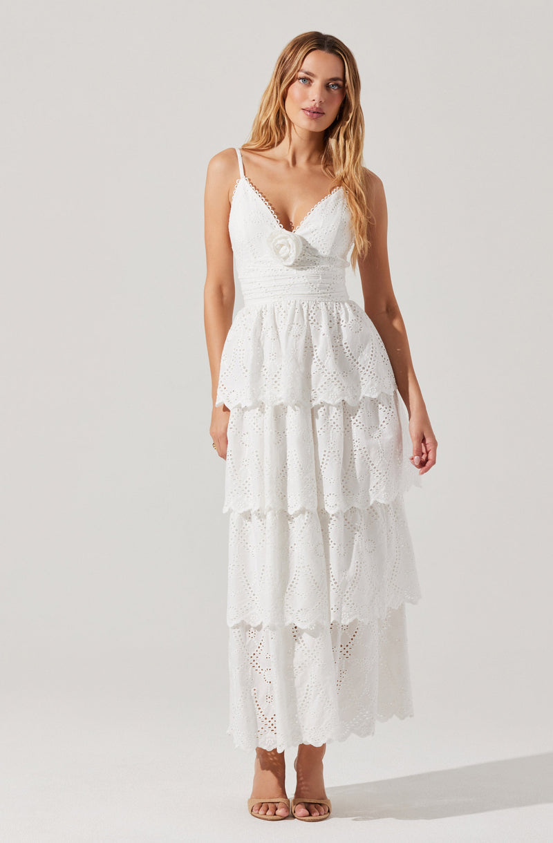 A woman is wearing a white sleeveless dress with a fitted bodice and thin spaghetti straps. The bodice features a decorative flower detail at the center. The skirt has multiple tiers of ruffled, eyelet fabric that create a layered, flowing look. She is also wearing beige open-toe high heel sandals.