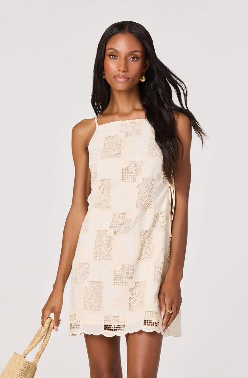 Woman wearing a cream-colored, patterned, sleeveless summer dress with delicate fabric and scalloped hem.