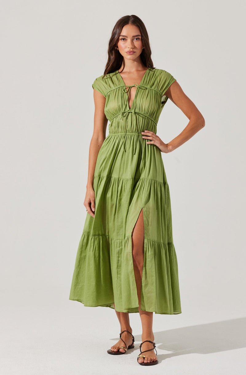 A woman is wearing a light green, tiered maxi dress with a gathered waist. The dress features short, puffed sleeves and a V-neckline with a drawstring tie at the front. It has a high slit on the left side, revealing her leg. She is also wearing brown strappy sandals.