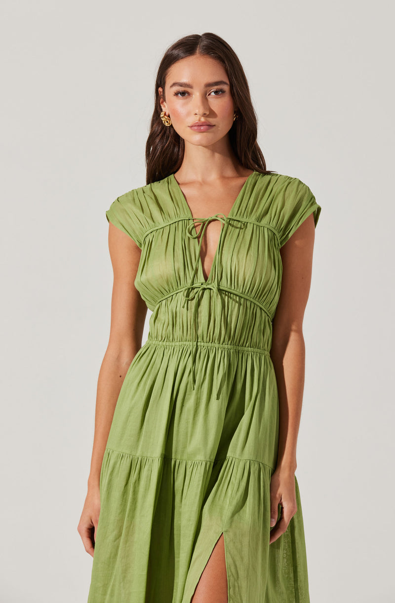 A woman is wearing a light green, sleeveless dress with a gathered, pleated design. The dress has a V-neckline with a small tie at the center, creating a keyhole opening. The fabric is gathered at the bust and waist, giving it a fitted appearance in those areas, and the skirt portion is loose and flowing with a slit on the side. The dress features multiple tiers, adding to its flowing, layered look.