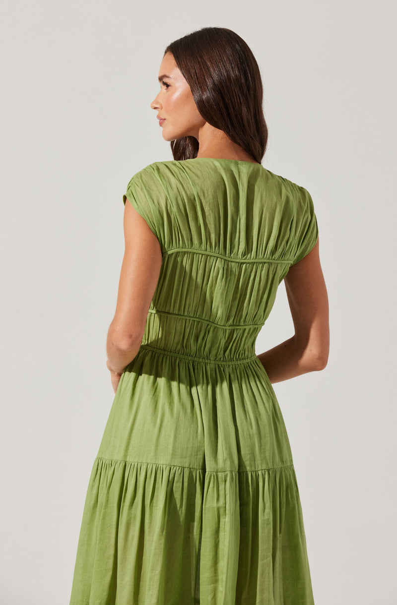 A woman is wearing a light green, tiered dress made of a soft, flowing fabric. The dress features short, gathered sleeves and multiple horizontal seams that create a gathered, textured effect throughout the garment. The waistline is slightly cinched, and the skirt section is gathered with a tiered design.