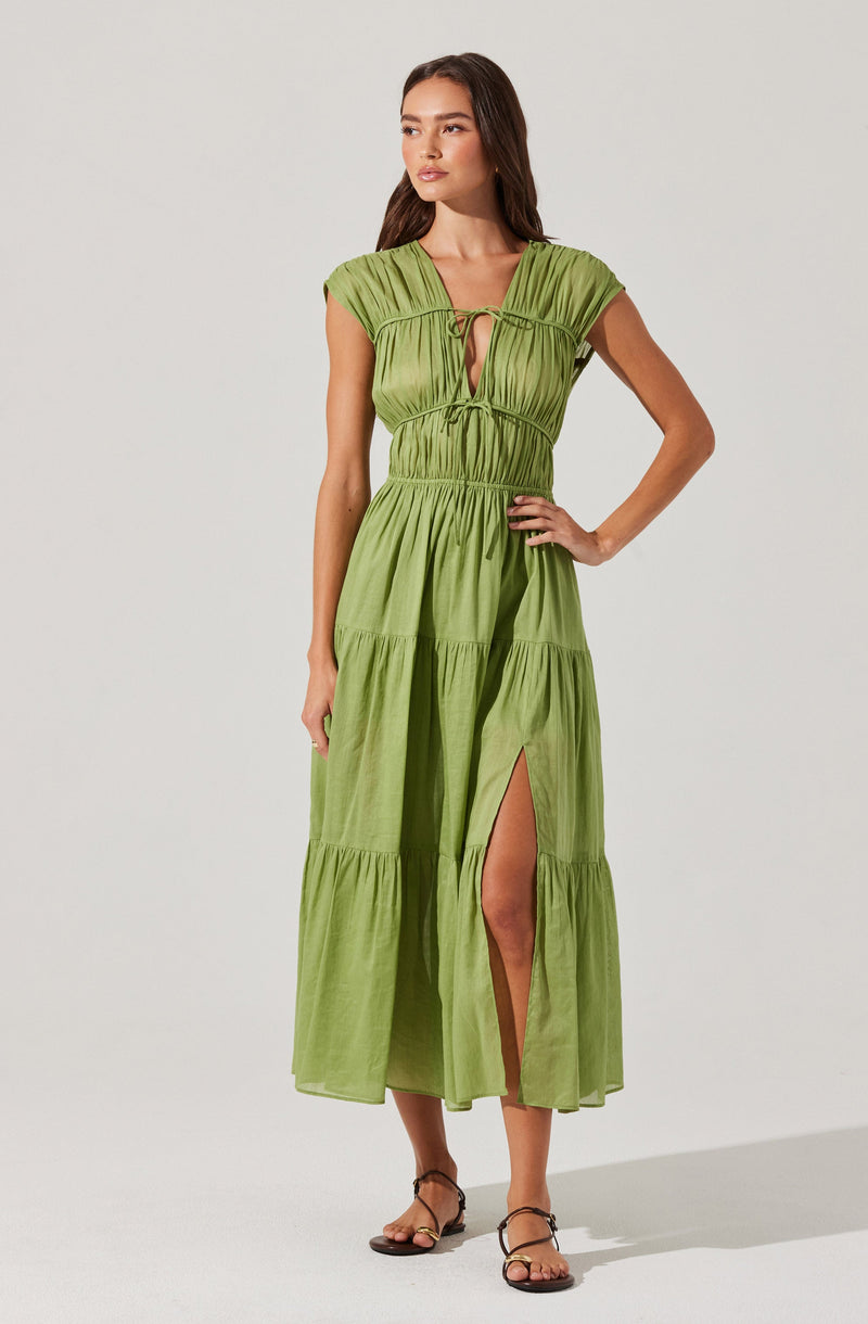 A woman is wearing a long, lime green dress with gathered details and a tiered skirt. The dress features a deep V neckline with a small keyhole and ties at the bust. It has short, cap sleeves and a high waistline. The skirt has a slit up to the knee, revealing her leg. She is also wearing strappy, flat sandals.