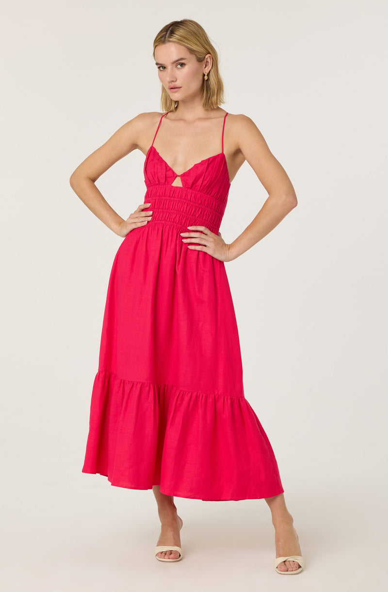 A woman is wearing a sleeveless, pink, spaghetti strap dress with a fitted bodice that features ruching and a small cutout detail near the neckline. The skirt is flowy and reaches down to her ankles with a tiered ruffle at the bottom. She is also wearing beige, open-toe heels.