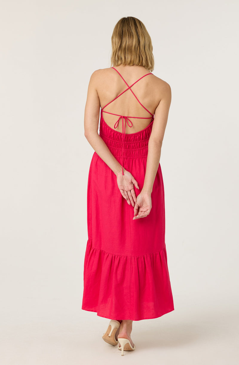A woman is wearing a sleeveless, floor-length dress in a bright pink color with a gathered waistband and a ruffled hem. The dress features thin spaghetti straps that crisscross on the back and tie at the waist. She is also wearing nude high heels.