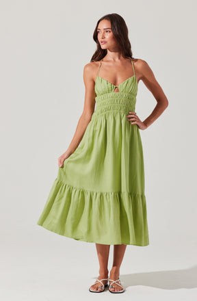 A woman is wearing a light green, sleeveless sundress with thin spaghetti straps. The dress features a ruched bodice with a small keyhole cutout at the center, and an elastic waistband. The skirt is ankle-length with a ruffled hem. She is also wearing flat sandals with thin white straps.