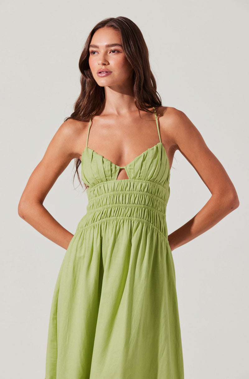 A woman is wearing a light green sundress with thin spaghetti straps. The top of the dress features gathered fabric and a small keyhole cutout below the bust. The waistline appears to be elasticized with horizontal ruched detailing, and the skirt is long and flowing with a gathered, full silhouette.