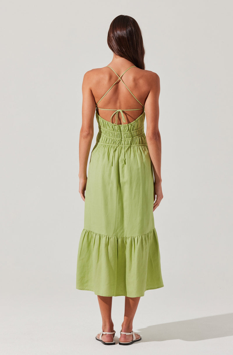 A woman is wearing a light green, sleeveless, backless maxi dress with thin spaghetti straps that crisscross at the back and tie in a bow. The dress features an elastic waistband and a tiered skirt with gathered fabric. She is also wearing flat sandals.