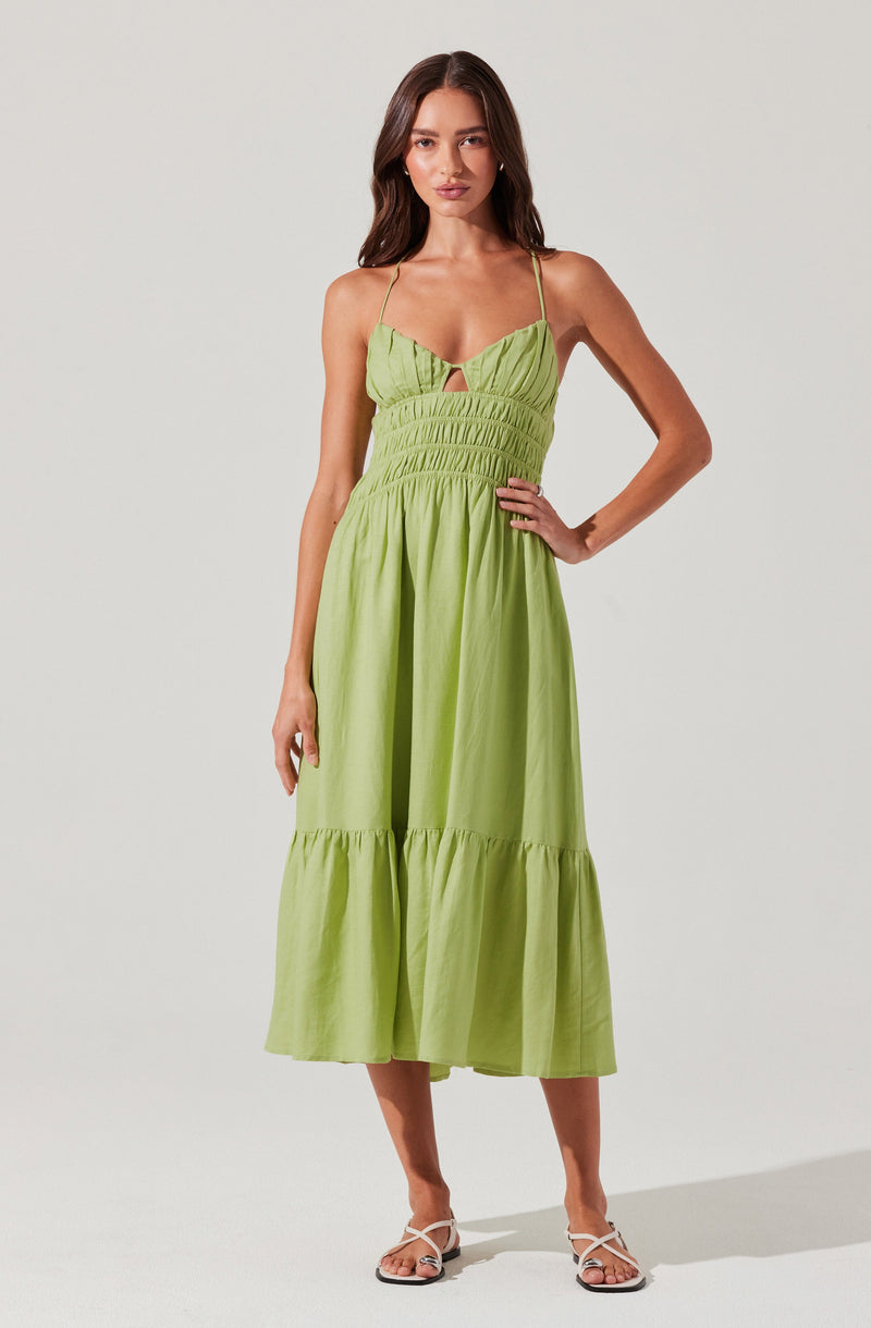A woman is wearing a light green, sleeveless maxi dress with thin spaghetti straps. The dress features a gathered, ruched bodice with a small keyhole cutout in the center, and a fitted waist. The skirt is voluminous with a tiered design, creating a flowing silhouette.