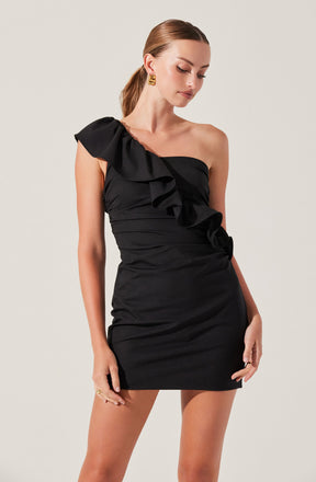 A woman is wearing a black, one-shoulder mini dress with ruffle details along the strap and down the front. The dress is fitted and has a slightly gathered design around the waist and hips. She accessorizes with gold earrings and rings.