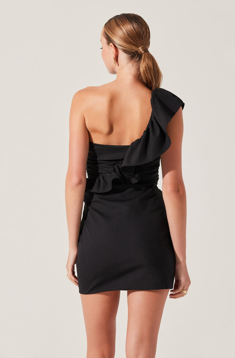 A woman is wearing a black, one-shoulder dress with a large ruffle detail on the shoulder and around the waist area. The dress is fitted and above knee-length.
