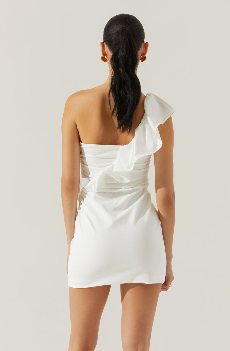 A woman is wearing a white, one-shoulder dress with a ruffle detail on the shoulder and gathered fabric at the bodice. The dress is fitted and short, ending above the knees. She has long, dark hair styled in a ponytail and is accessorizing with gold earrings.