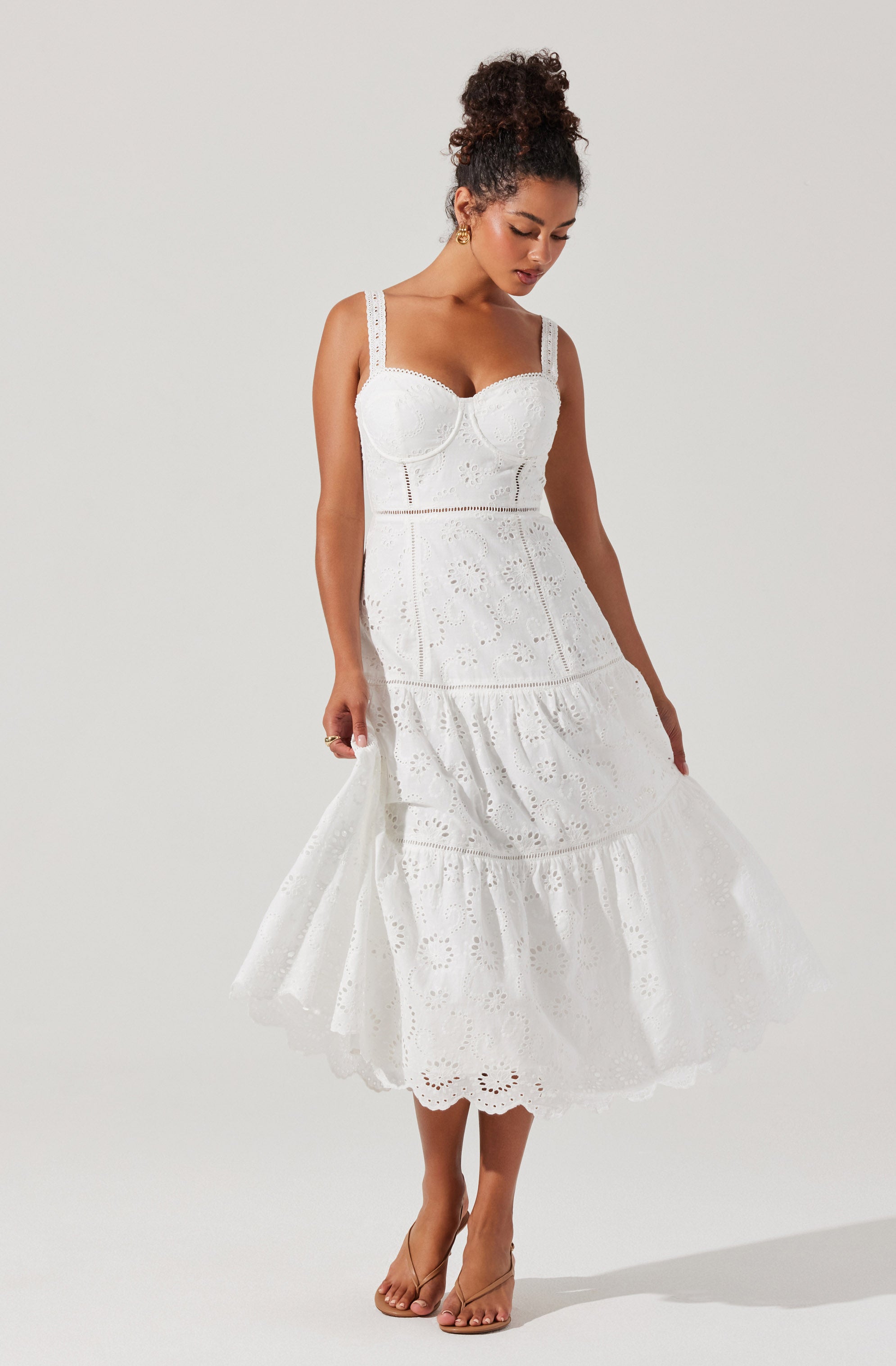 A woman is wearing a white, sleeveless, eyelet lace dress with wide straps. The bodice is fitted with a sweetheart neckline, and the skirt is tiered with a ruffled hem, creating a flowy silhouette. She is barefoot and has her hair styled in a high, curly bun.