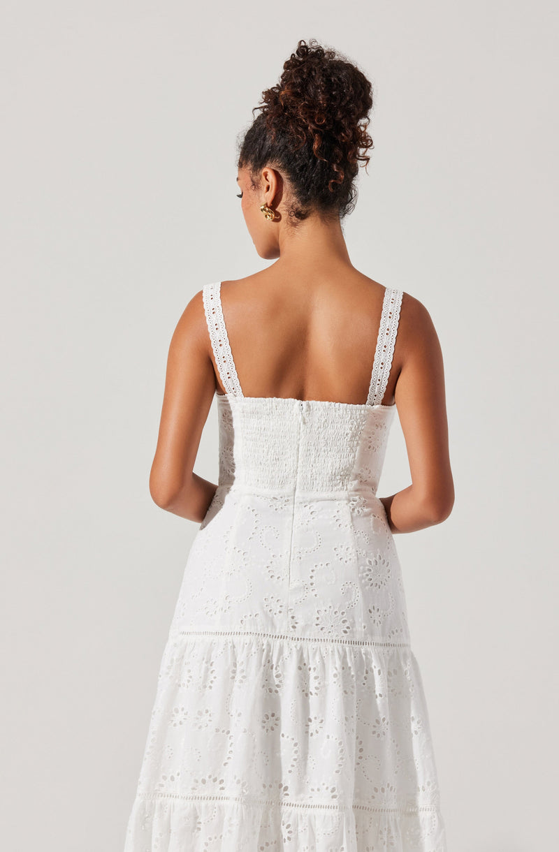 A woman is wearing a sleeveless white dress with embroidered eyelet details. The dress has wide straps with lace trim, a fitted bodice, and a flowing skirt with multiple tiers.