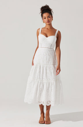 A woman is wearing a white, sleeveless, eyelet lace dress with broad shoulder straps. The dress features a fitted bodice with a sweetheart neckline and a tiered, flowing skirt that falls to mid-calf length. The fabric has intricate embroidered eyelet patterns throughout.