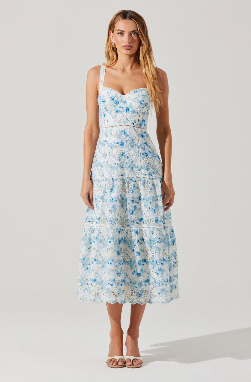 A woman is wearing a sleeveless, ankle-length dress with a fitted bodice and a full skirt. The dress features a white base with a blue floral pattern throughout, including on the wide shoulder straps, bodice, and skirt. The fabric appears lightweight and textured, with a tiered design on the skirt and delicate lace or embroidery details. She is also wearing nude-colored open-toe sandals.