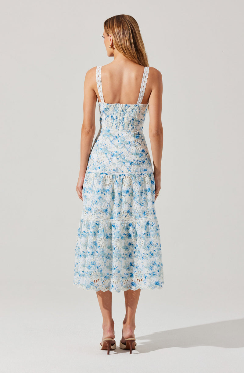 A woman is wearing a sleeveless, white dress with wide straps. The dress features a blue floral pattern with lace detailing on the straps, bodice, and hem. The skirt is tiered and midi-length, creating a flowy silhouette. She is also wearing beige high-heeled shoes.