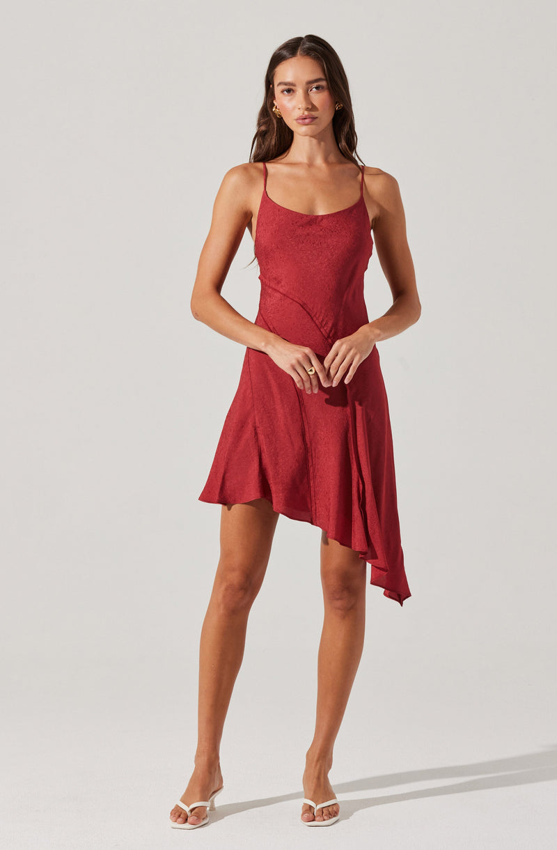 A woman is wearing a sleeveless, deep red dress with thin spaghetti straps. The dress has a slight shimmer and features a draped, asymmetrical hemline on one side, creating a flowing and elegant silhouette.