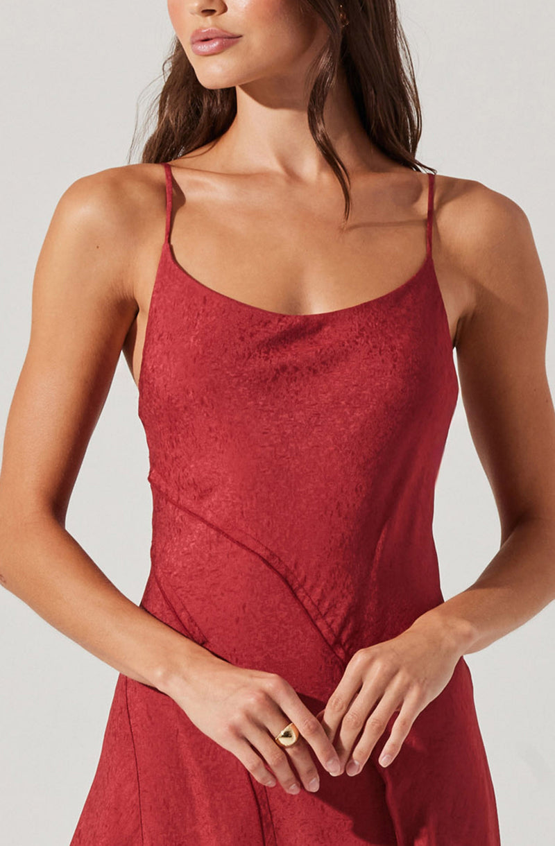 A woman is wearing a sleeveless, red slip dress with thin spaghetti straps. The fabric has a slightly textured appearance, and the dress features subtle, curved seam details across the waist.