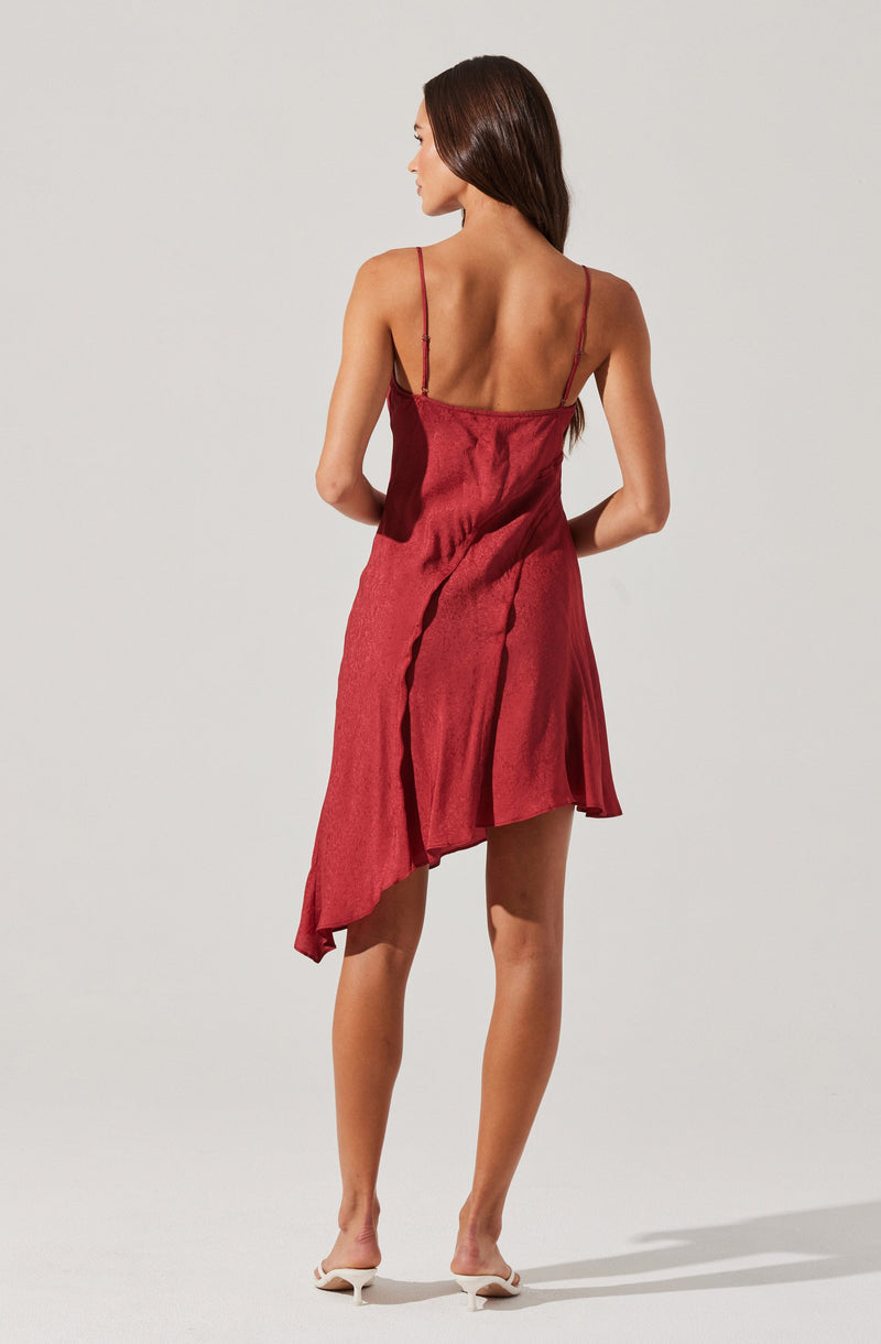 A woman is wearing a sleeveless, red dress with thin spaghetti straps. The dress has an asymmetrical hemline that is longer on one side, creating a flowing, layered appearance. The fabric appears to be silky with a slight sheen. She pairs the dress with white heeled shoes.